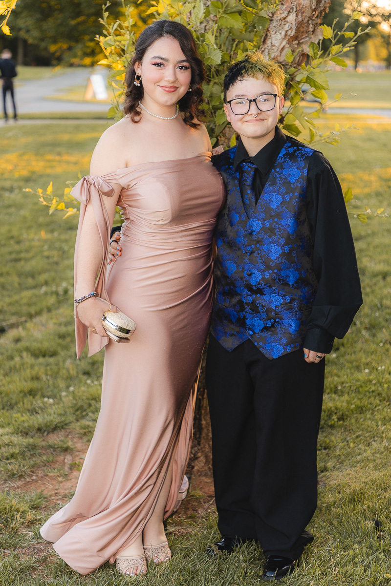 Two young people posing outdoors in formal wear under a tree: a woman in a blush off-the-shoulder gown with a pearl necklace and clutch, and a person in a blue floral vest with a black shirt and glasses.