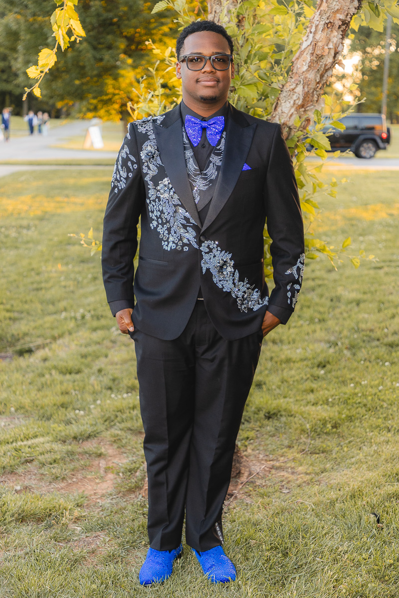 Man in a black suit with blue embroidered floral pattern, blue bow tie, and blue shoes posing outdoors by a tree in a park.”," Formal portrait of a man in a black suit with ornate silver-blue embroidery, bright blue bow tie, and matching blue shoes, standing with hands in pockets."," A well-dressed man wearing a tuxedo with floral embroidery, blue accessories, and sunglasses, standing in a grassy park." ," Individual in a stylish black suit with decorative blue embroidery and matching accessories posing by a tree in a sunny park."," Young man in a black tuxedo with floral embroidery, blue bow tie, and cobalt shoes, outdoors on a fall day."]} }```**Note**: The JSON has a single key