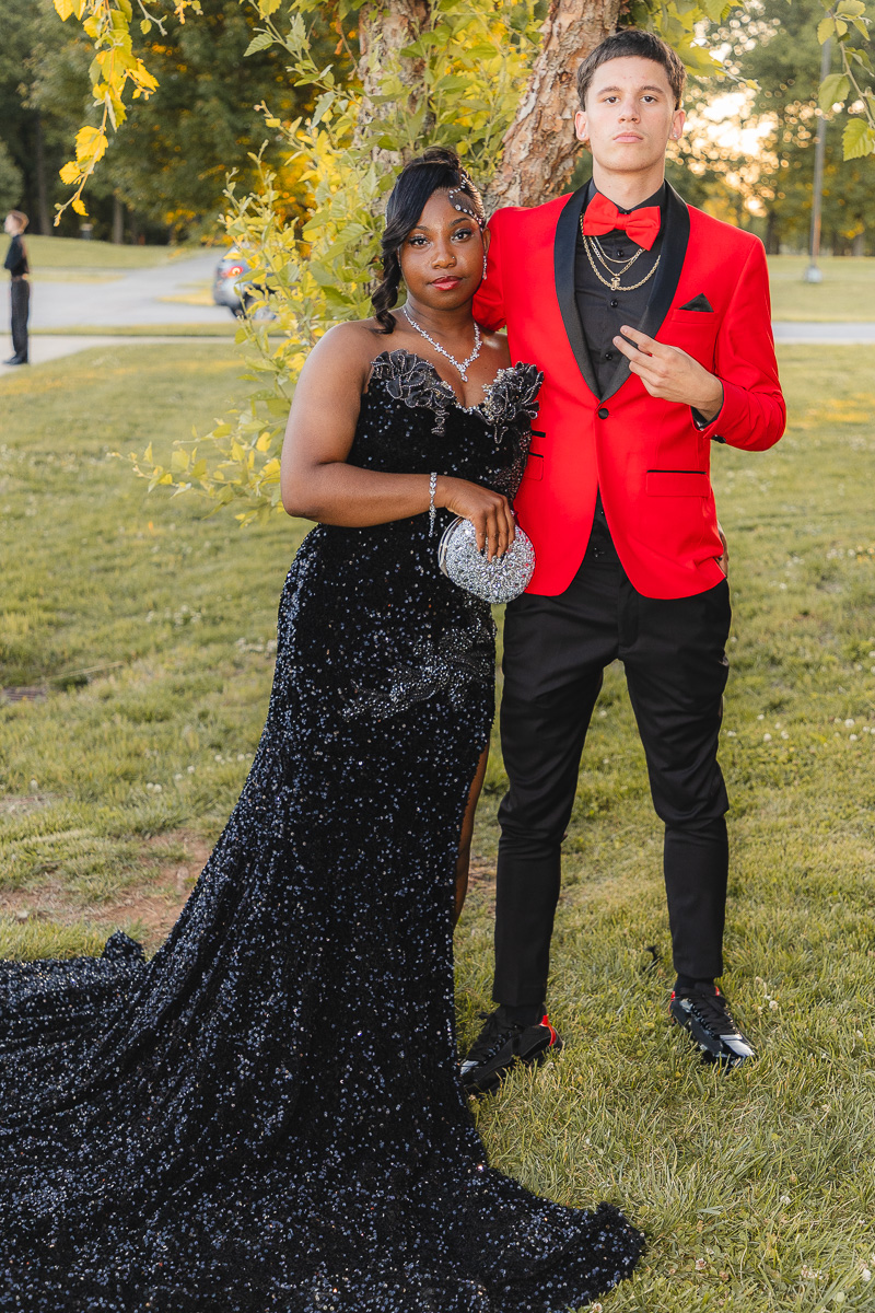 Couple in formal attire posing outdoors; woman in a black sequin gown with a glitter clutch, man in a red suit jacket and bow tie.