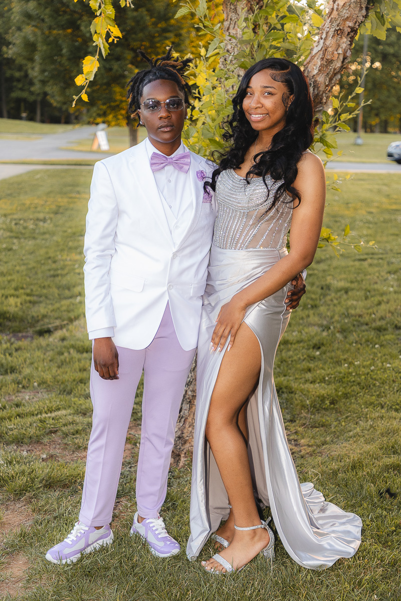 Two young people pose together outdoors in formal wear: a person in a white suit with lavender pants and bow tie beside a woman in a silver gown with a high slit, both smiling under a tree.