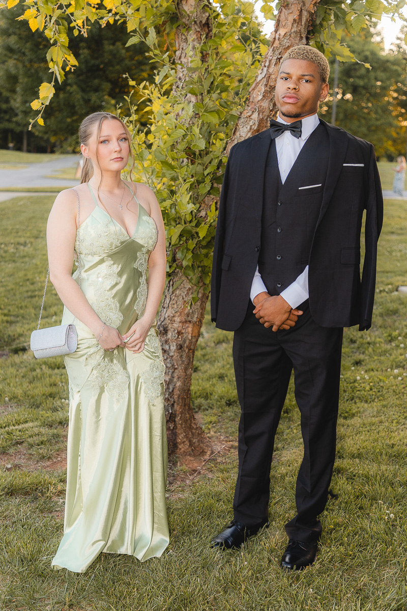 Two teens in formal wear stand beside a tree on a grassy field, the girl in a pale green gown and the boy in a black tuxedo with a bow tie, posing for a photo.