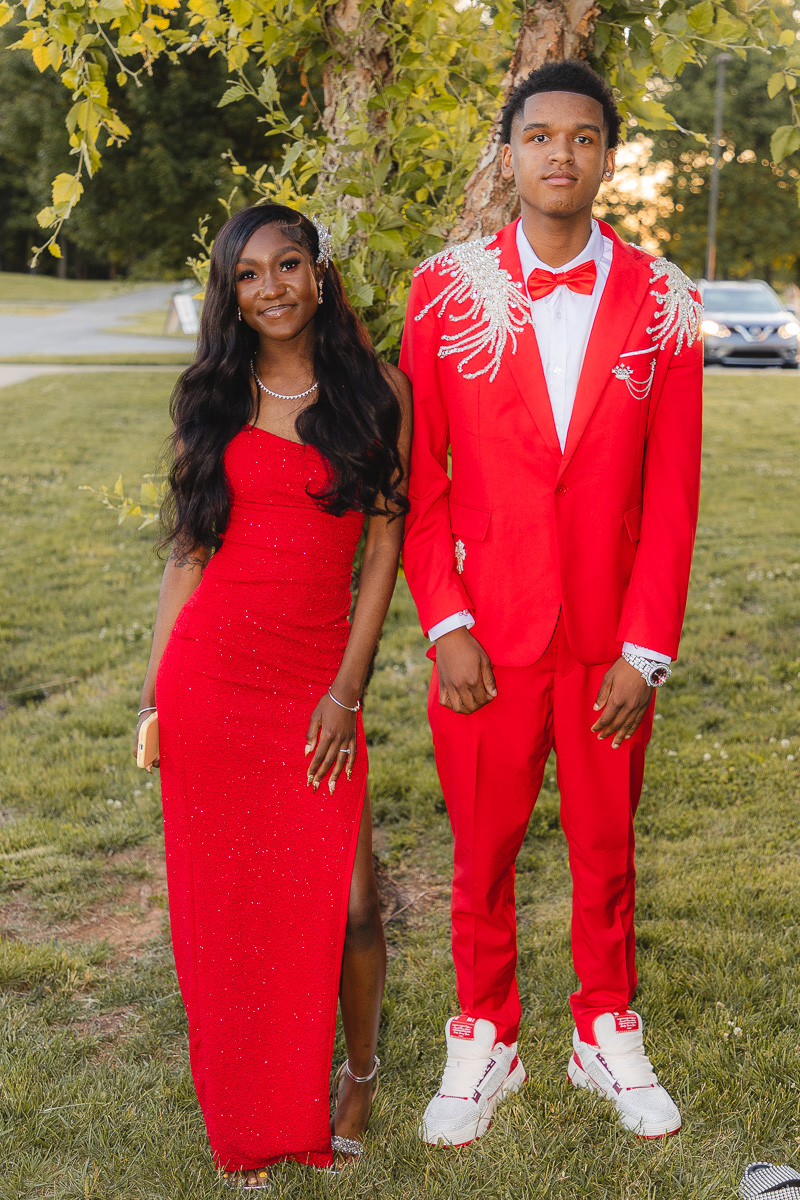 Two teens in red formal wear posing outdoors under a tree: the girl in a sparkling red gown and the boy in a red suit with a white shirt and bow tie.