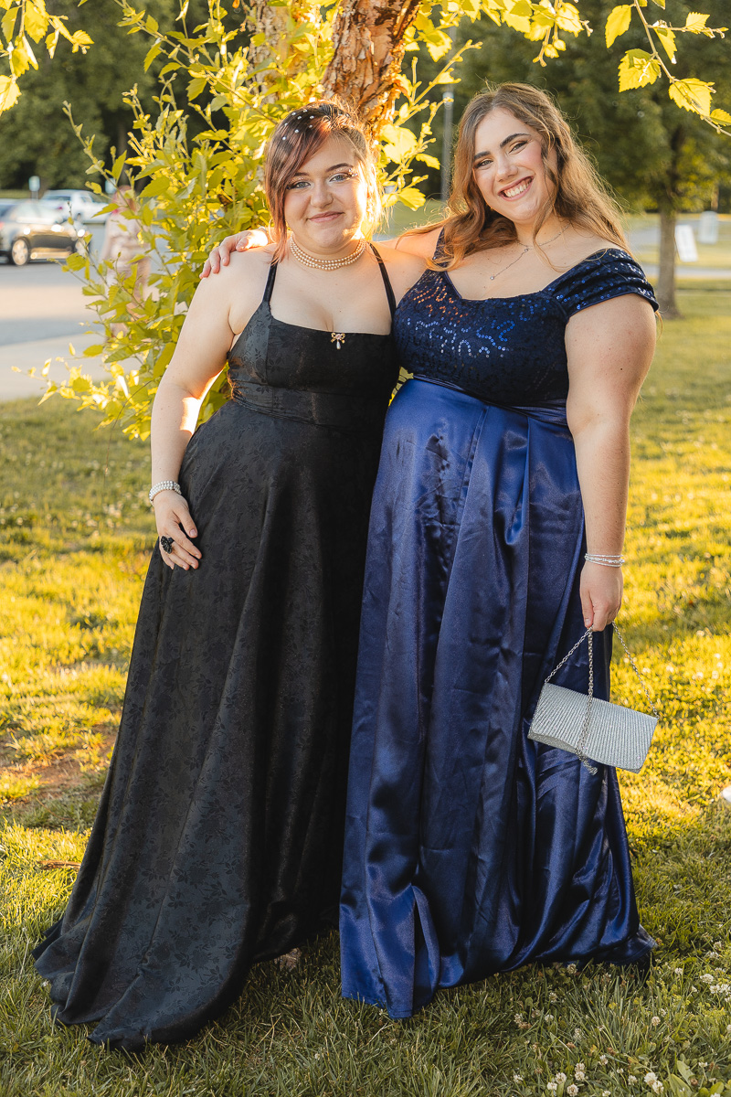 Two young women in formal gowns posing outdoors by a tree, smiling at the camera; left in a black dress, right in a blue satin dress with a silver clutch.