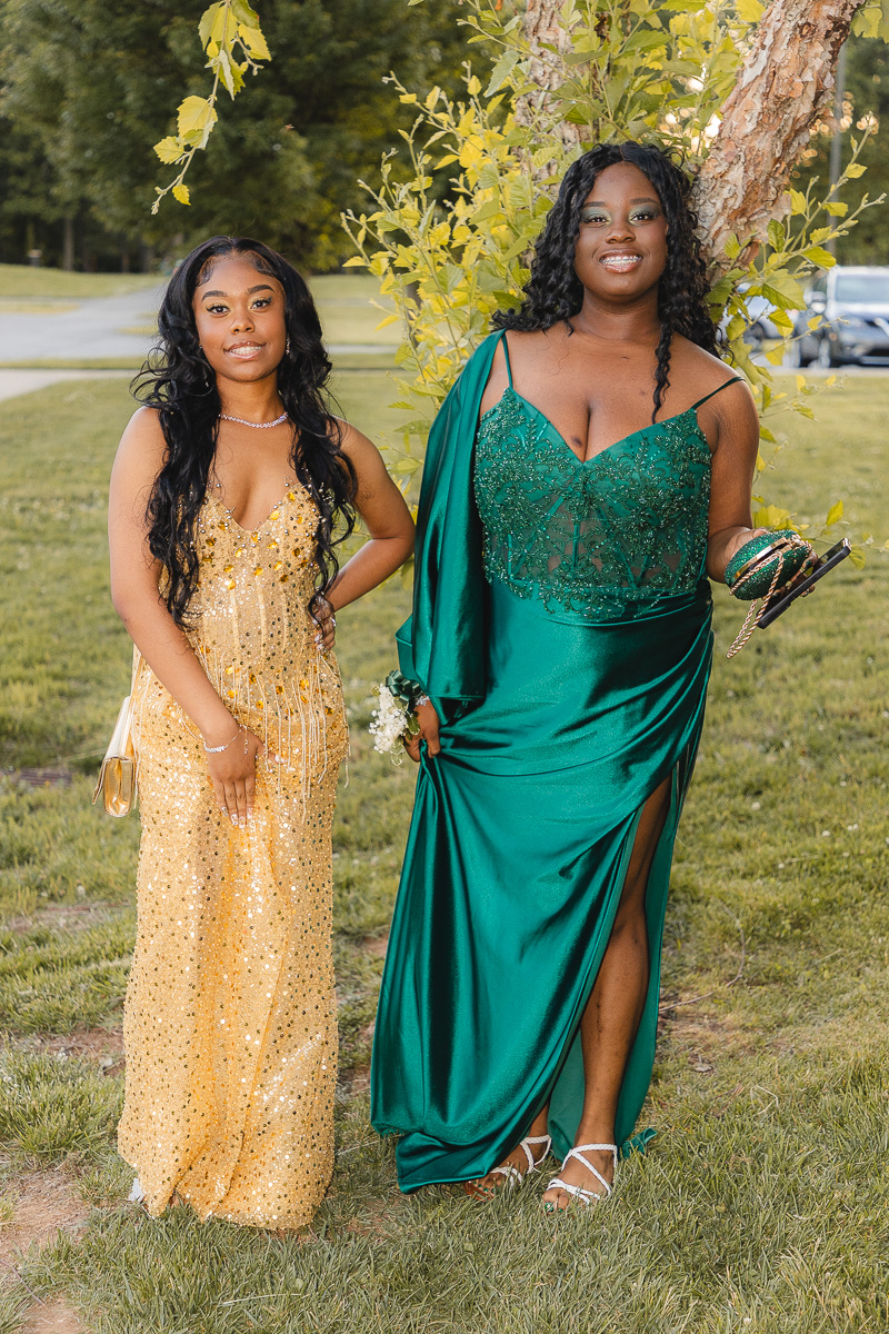 Two young women in formal gowns posing outdoors on a grassy area, one in a gold sequin dress and the other in a green emerald gown, with a tree behind them.