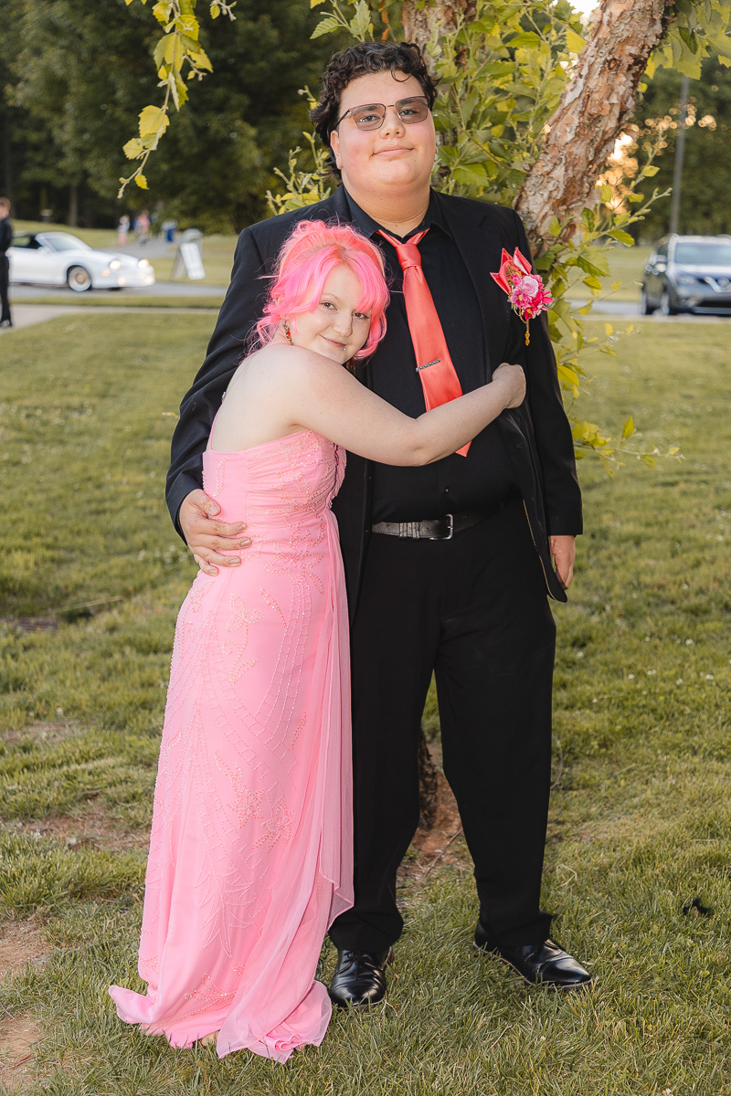 Two friends in formal attire posing outdoors by a tree; one in a pink gown with pink hair hugs the other in a black suit with a red tie and pink boutonniere, smiling at the camera.