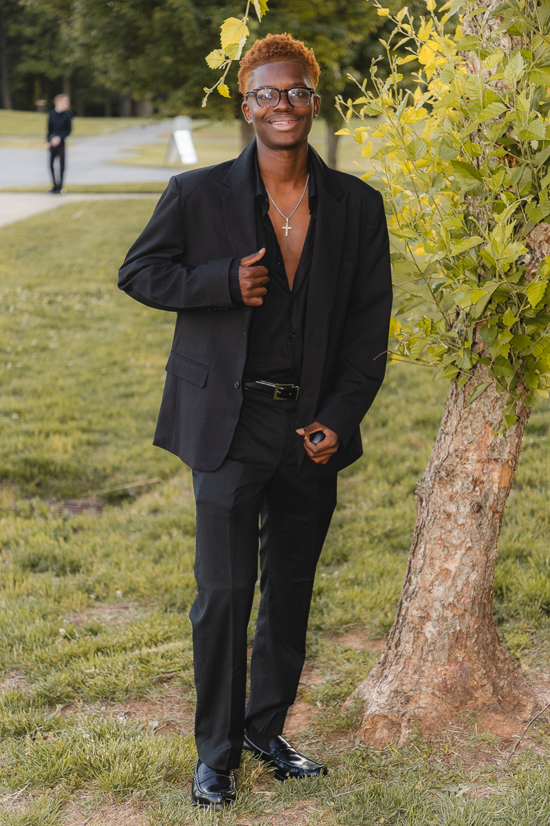 Young man in a black suit adjusts his lapel and smiles at the camera beside a tree in a park.