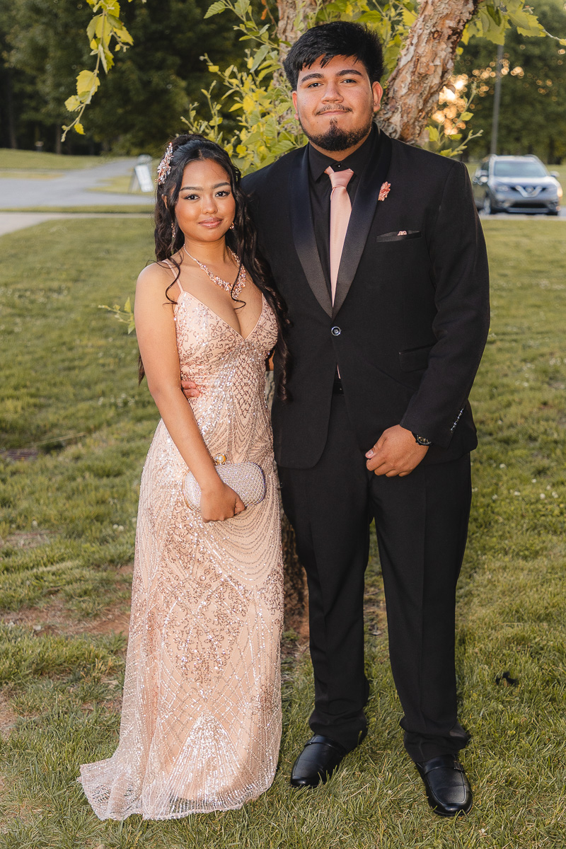 Couple standing together outdoors in formal attire: woman in a champagne sequin gown holding a small clutch and necklace, man in a black suit with a pink tie, both smiling near a tree on a grassy area.