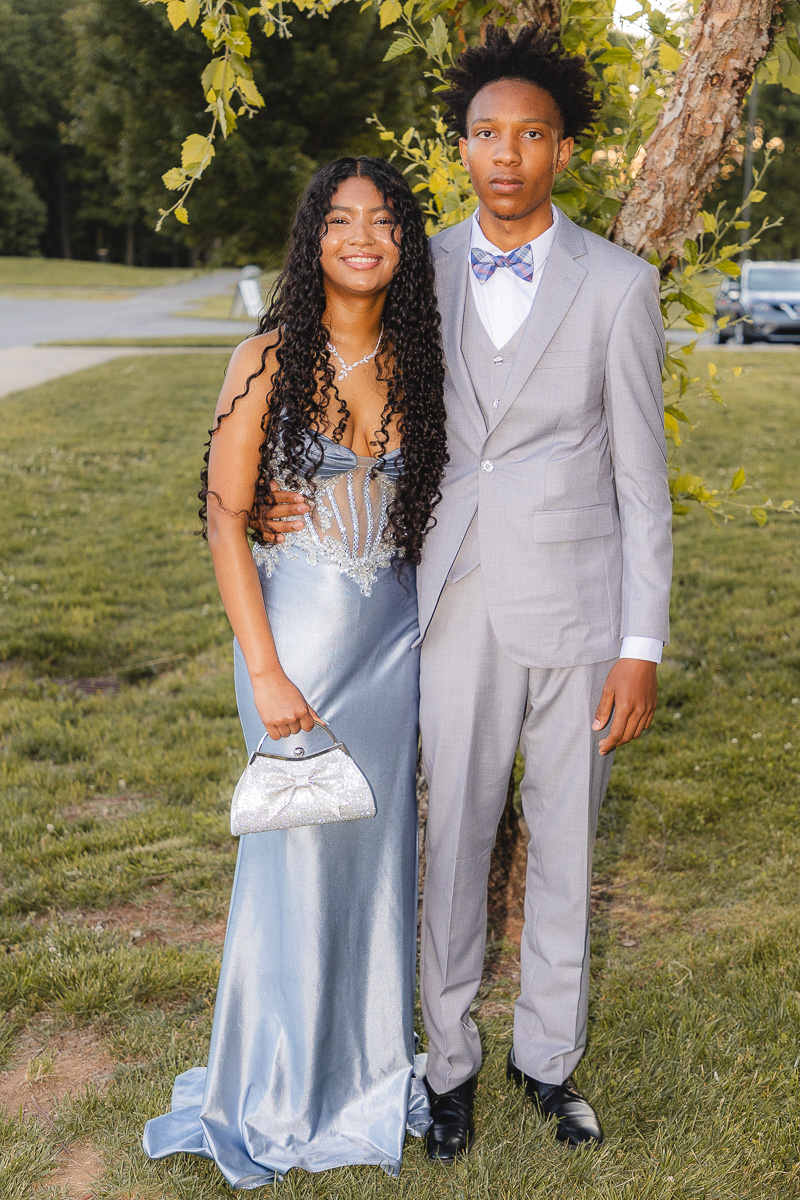 Teen couple posing outdoors in formal wear: girl in a light blue gown with a silver clutch, boy in a gray suit.