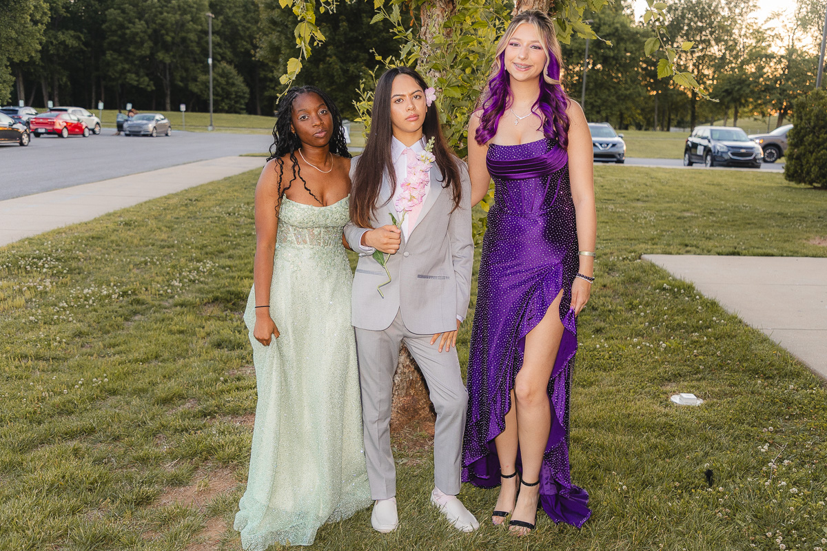 Three teens in formal wear posing on a grassy area; mint green dress, gray suit with a pink flower, and a purple gown with a slit.