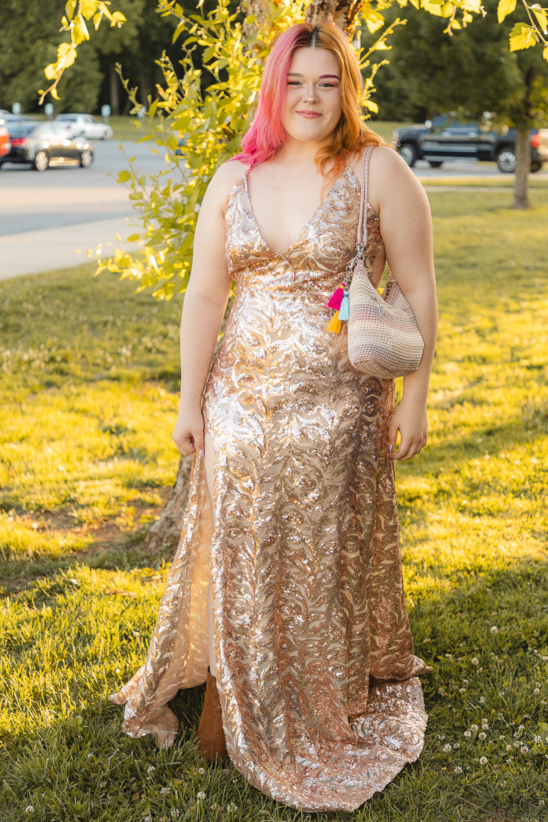 Woman in a champagne sequin gown standing on grass outdoors, carrying a beige woven purse with colorful tassels.