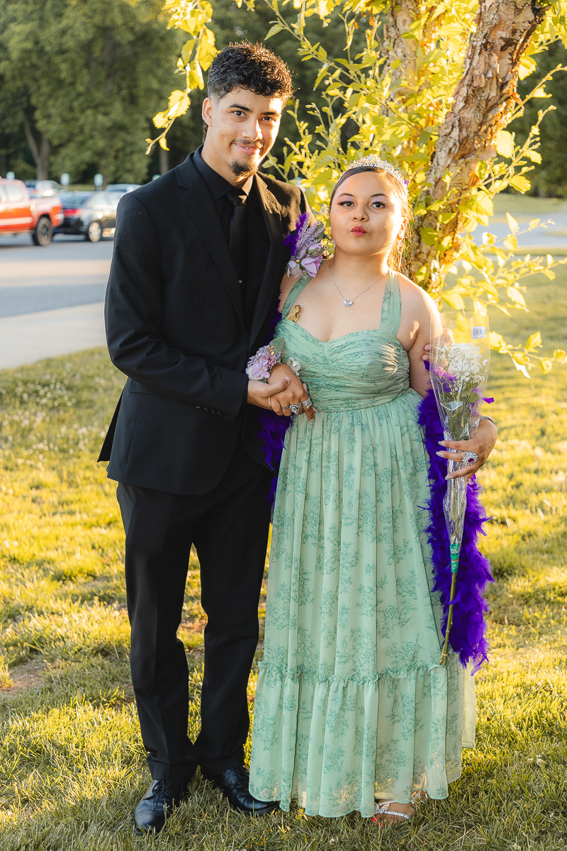 A smiling couple dressed formally outdoors: man in a black suit holds a woman in a mint green gown with flowers and a purple feather boa.