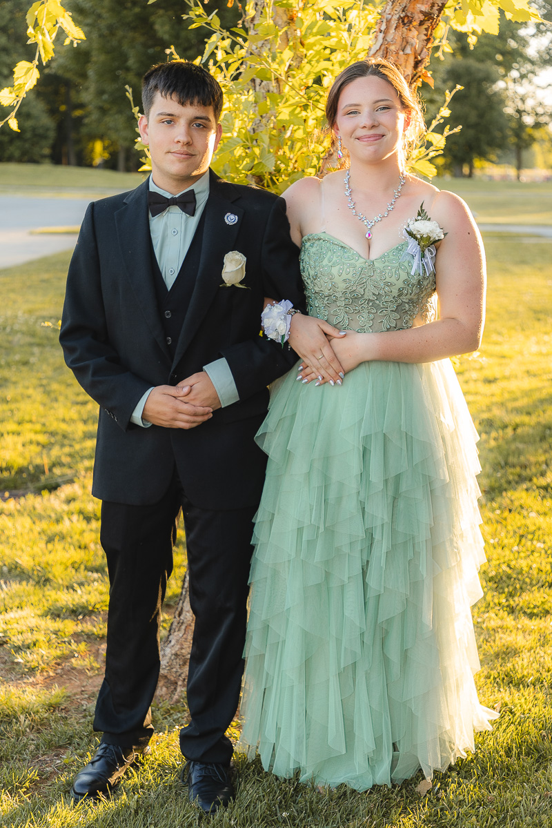A young couple outdoors in formal wear, arms linked; the boy in a black suit and bow tie, the girl in a mint green layered gown with a statement necklace.