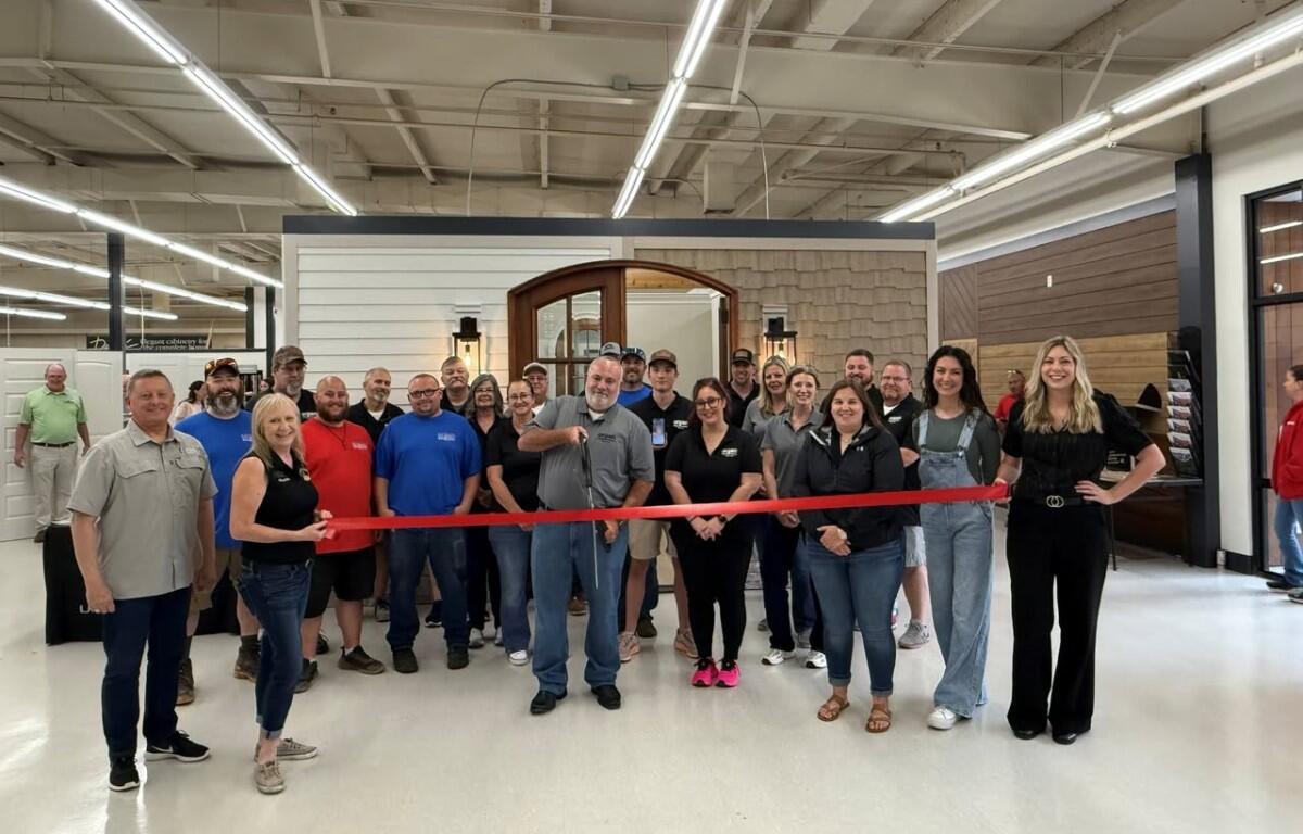 Group of coworkers gathered for a ribbon-cutting ceremony in a showroom, standing behind a stretched red ribbon in front of a doorway display.
