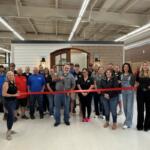 Group of coworkers gathered for a ribbon-cutting ceremony in a showroom, standing behind a stretched red ribbon in front of a doorway display.