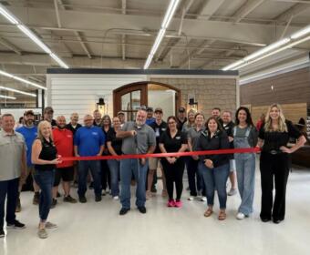 Group of coworkers gathered for a ribbon-cutting ceremony in a showroom, standing behind a stretched red ribbon in front of a doorway display.