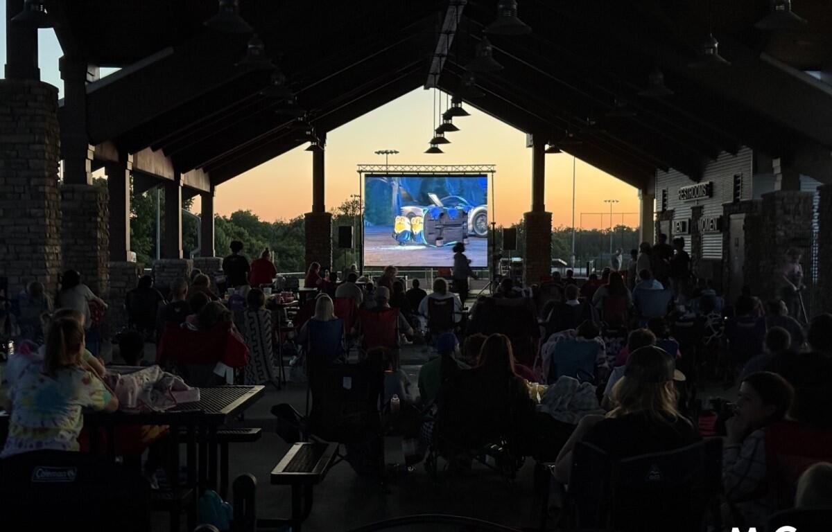 Audience seated under a pavilion watching a large outdoor movie screen at sunset.