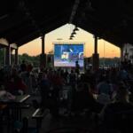 Audience seated under a pavilion watching a large outdoor movie screen at sunset.