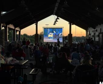 Audience seated under a pavilion watching a large outdoor movie screen at sunset.