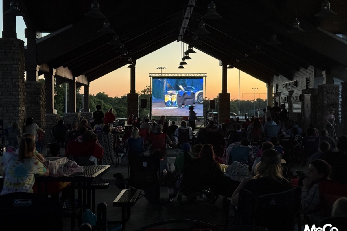 Audience seated under a pavilion watching a large outdoor movie screen at sunset.
