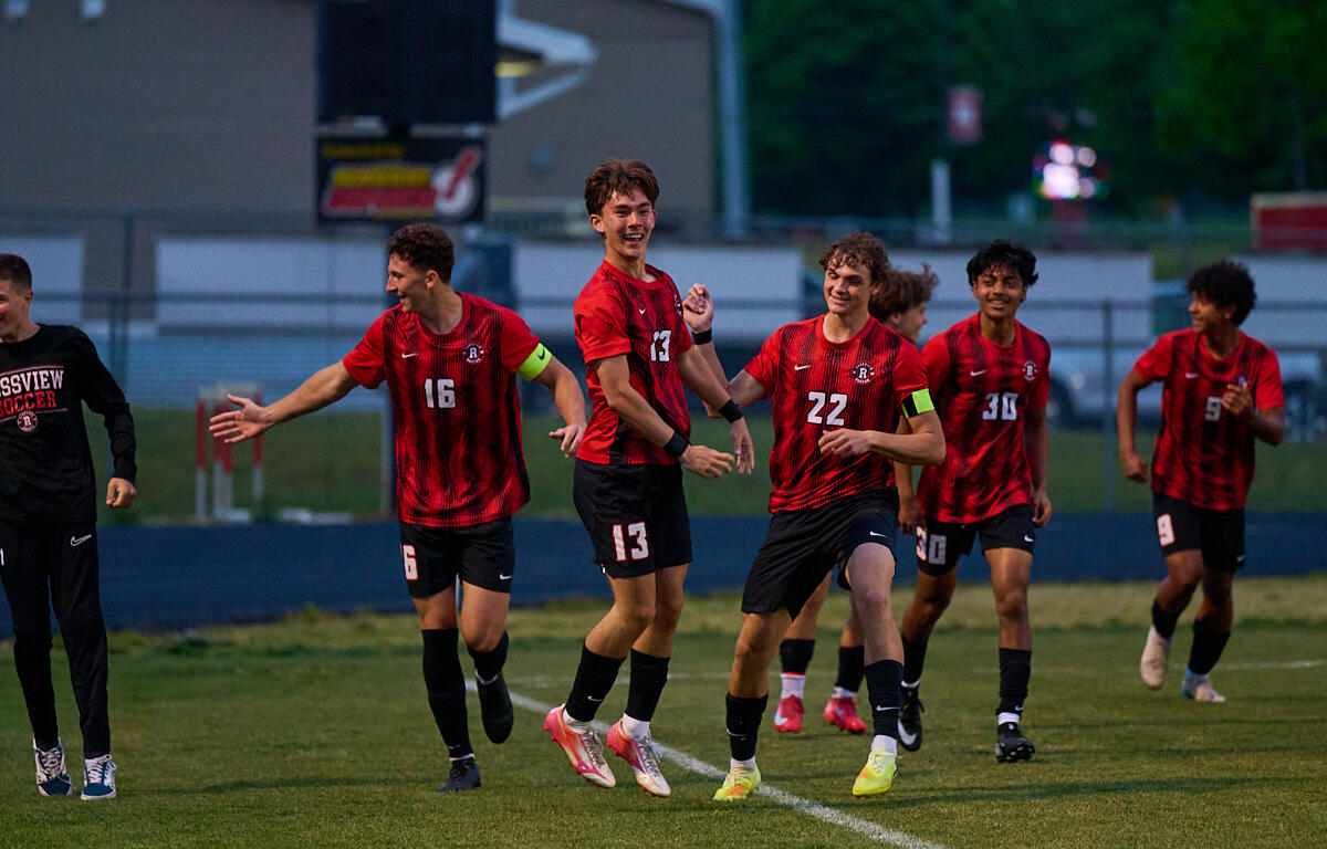 Group of young soccer players in red and black uniforms celebrating on a grassy field after a game.