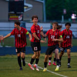 Group of young soccer players in red and black uniforms celebrating on a grassy field after a game.