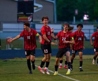 Group of young soccer players in red and black uniforms celebrating on a grassy field after a game.