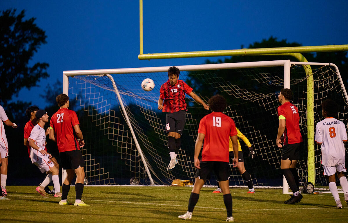 Soccer game at dusk; a red-jersey player jumps to head the ball near the goal while teammates watch.
