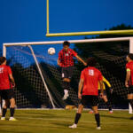 Soccer game at dusk; a red-jersey player jumps to head the ball near the goal while teammates watch.
