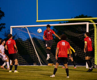 Soccer game at dusk; a red-jersey player jumps to head the ball near the goal while teammates watch.