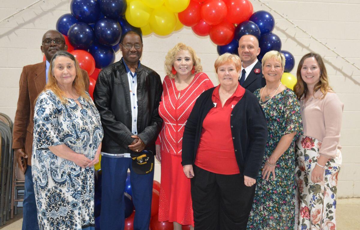 Group of diverse adults posing together under a red, yellow, and blue balloon arch at an indoor event, smiling for the photo.