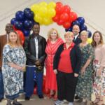 Group of diverse adults posing together under a red, yellow, and blue balloon arch at an indoor event, smiling for the photo.
