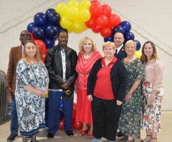 Group of diverse adults posing together under a red, yellow, and blue balloon arch at an indoor event, smiling for the photo.
