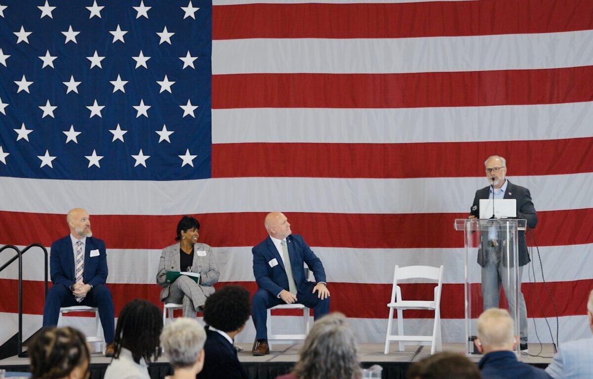 Panel discussion on stage with a large U.S. flag backdrop; four people seated, one at a clear podium speaking into a microphone.