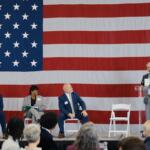 Panel discussion on stage with a large U.S. flag backdrop; four people seated, one at a clear podium speaking into a microphone.