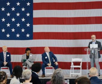 Panel discussion on stage with a large U.S. flag backdrop; four people seated, one at a clear podium speaking into a microphone.