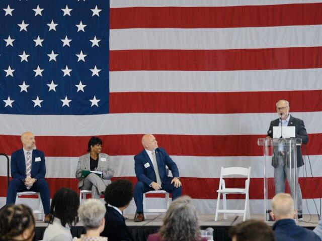 Panel discussion on stage with a large U.S. flag backdrop; four people seated, one at a clear podium speaking into a microphone.
