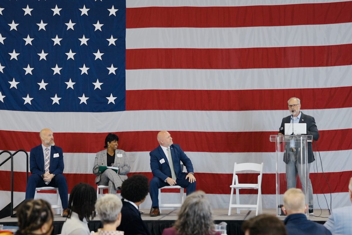 Panel discussion on stage with a large U.S. flag backdrop; four people seated, one at a clear podium speaking into a microphone.