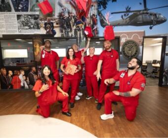 Group of seven people in red scrubs pose celebrating in a hallway with a helicopter mural behind them named and a circular emblem on the wall.