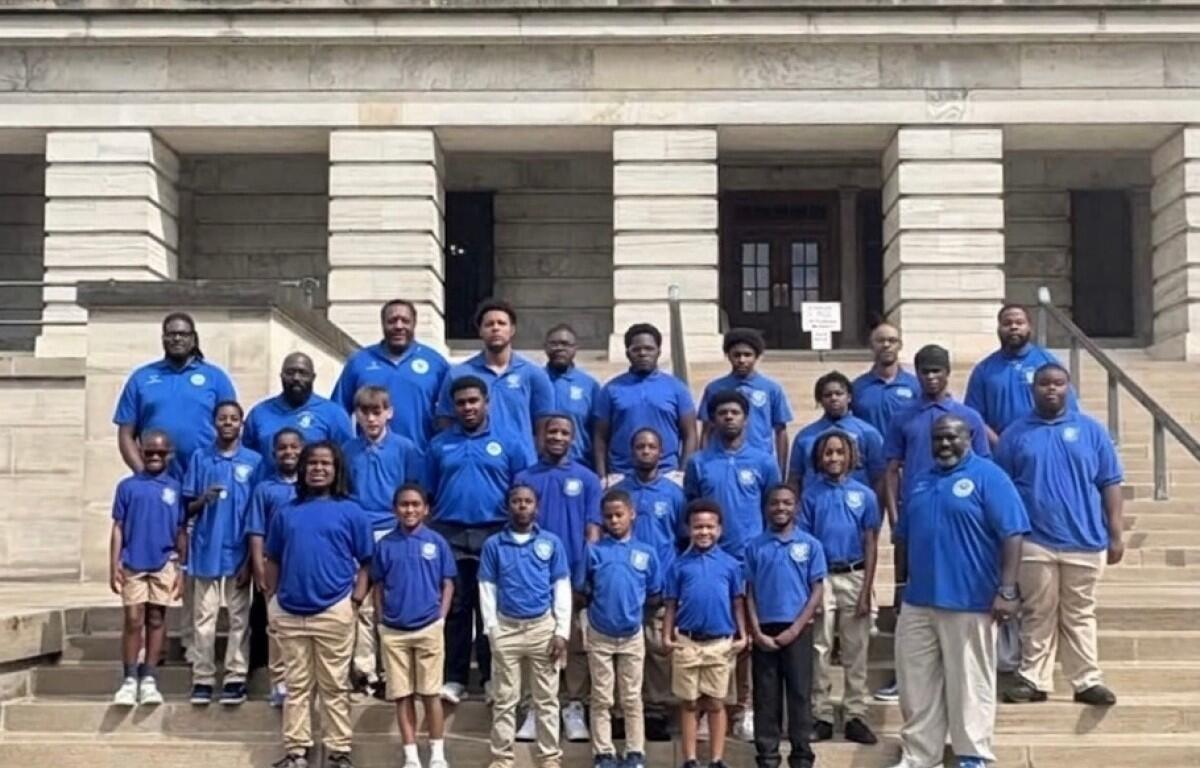 Group of people of varying ages wearing blue polo shirts posing on the stone steps of a large building. Photo resembles a team or class group portrait.