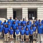 Group of people of varying ages wearing blue polo shirts posing on the stone steps of a large building. Photo resembles a team or class group portrait.