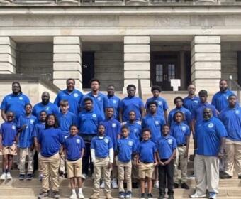 Group of people of varying ages wearing blue polo shirts posing on the stone steps of a large building. Photo resembles a team or class group portrait.
