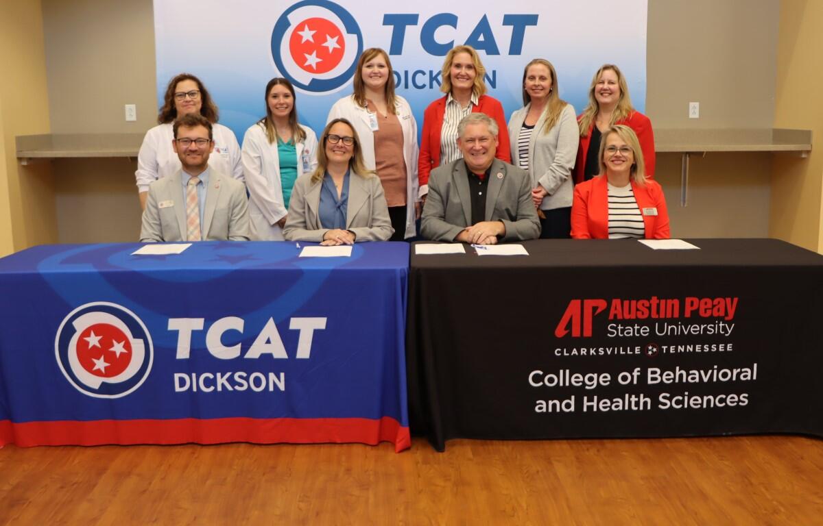 Group of ten professionals at a signing event, seated and standing behind two tables with TCAT Dickson and APSU banners in a classroom setting.