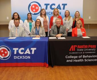 Group of ten professionals at a signing event, seated and standing behind two tables with TCAT Dickson and APSU banners in a classroom setting.