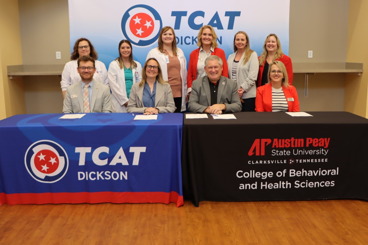 Group of ten professionals at a signing event, seated and standing behind two tables with TCAT Dickson and APSU banners in a classroom setting.