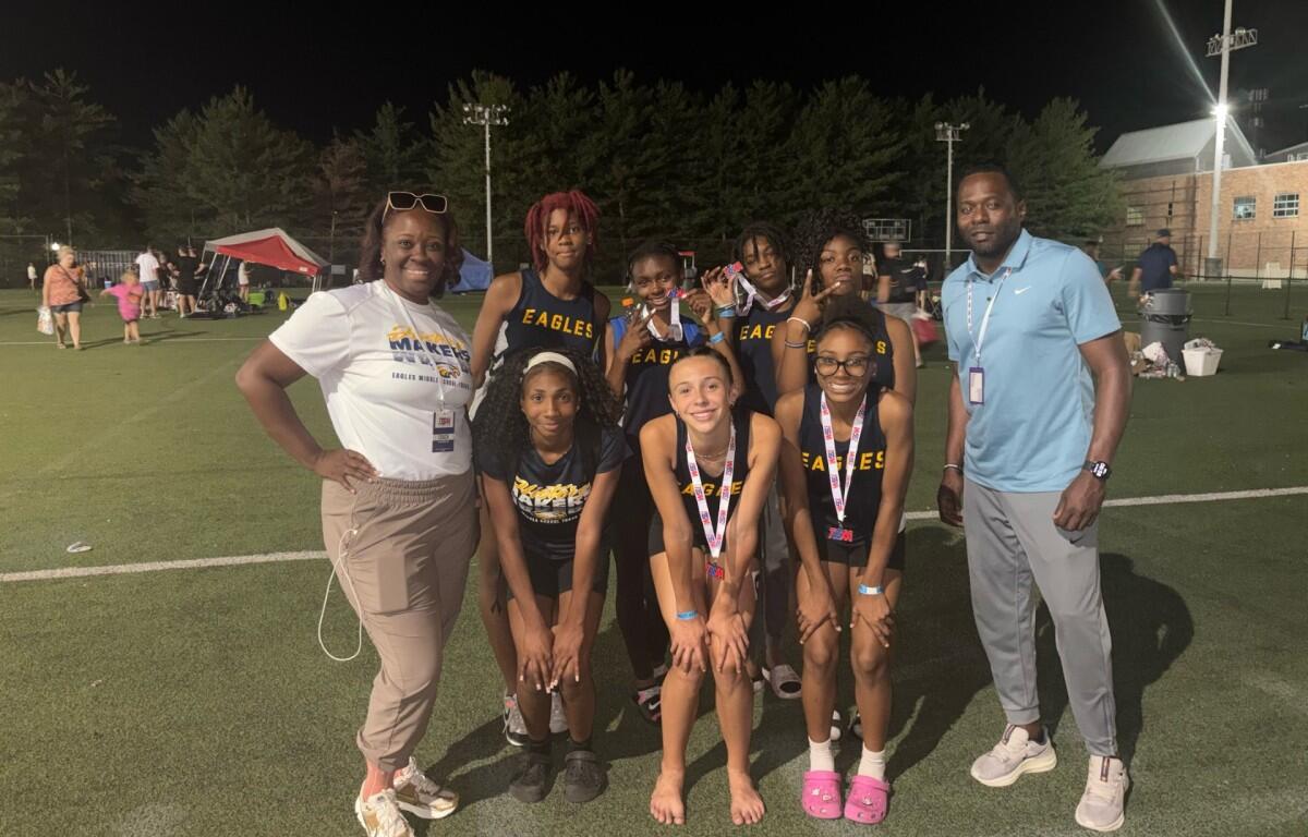 Group of young girls in Eagles uniforms posing with two adults on a lit outdoor field at night; medals around their necks.