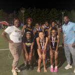 Group of young girls in Eagles uniforms posing with two adults on a lit outdoor field at night; medals around their necks.