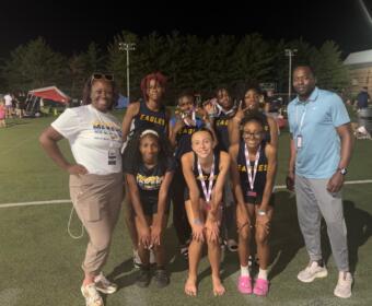 Group of young girls in Eagles uniforms posing with two adults on a lit outdoor field at night; medals around their necks.