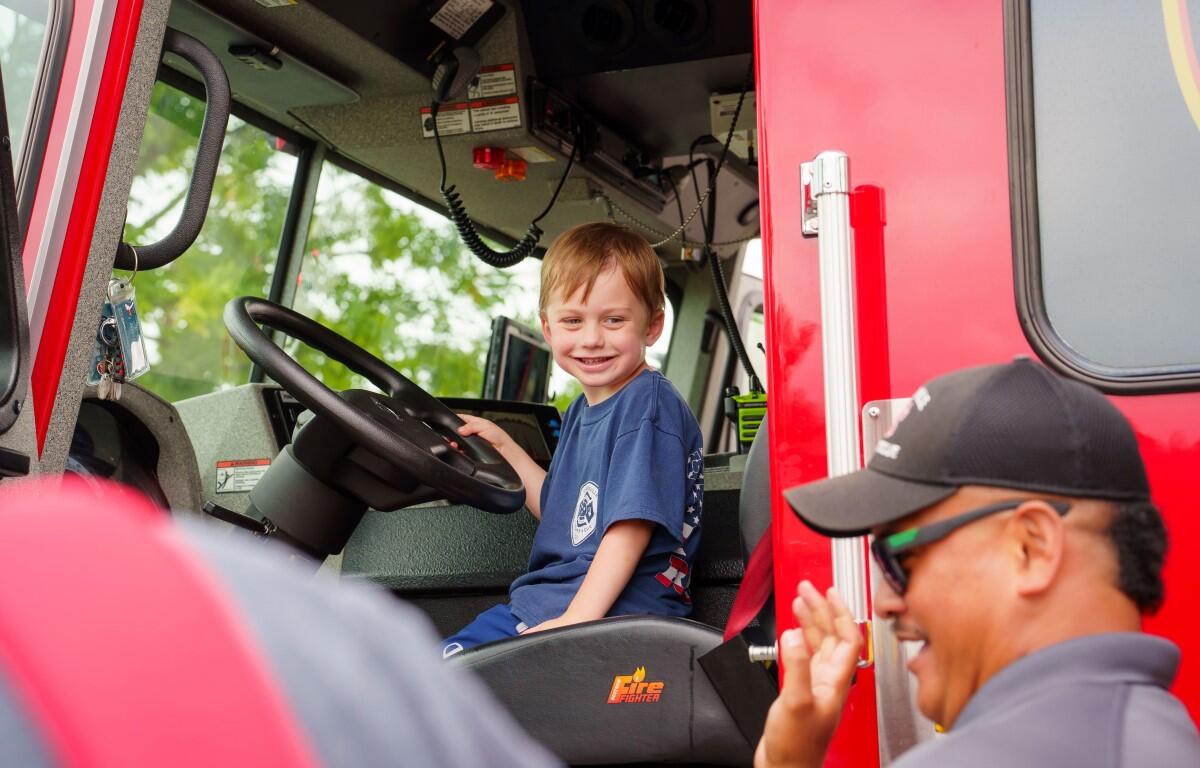 Young boy sits in the driver’s seat of a red fire truck, smiling as a firefighter waves from outside the cab.