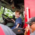 Young boy sits in the driver’s seat of a red fire truck, smiling as a firefighter waves from outside the cab.