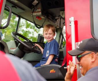 Young boy sits in the driver’s seat of a red fire truck, smiling as a firefighter waves from outside the cab.